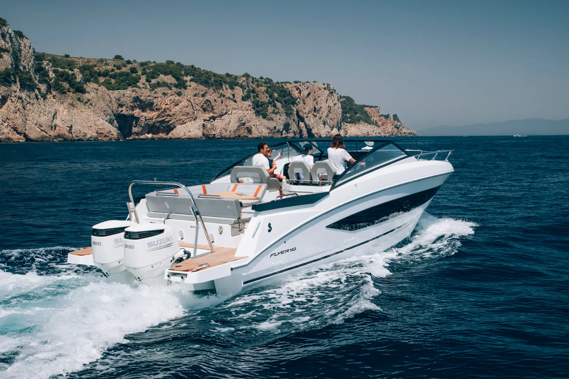 Vier mensen varen met een Beneteau Flyer 10 motorboot over helderblauw water langs een rotsachtige kustlijn.