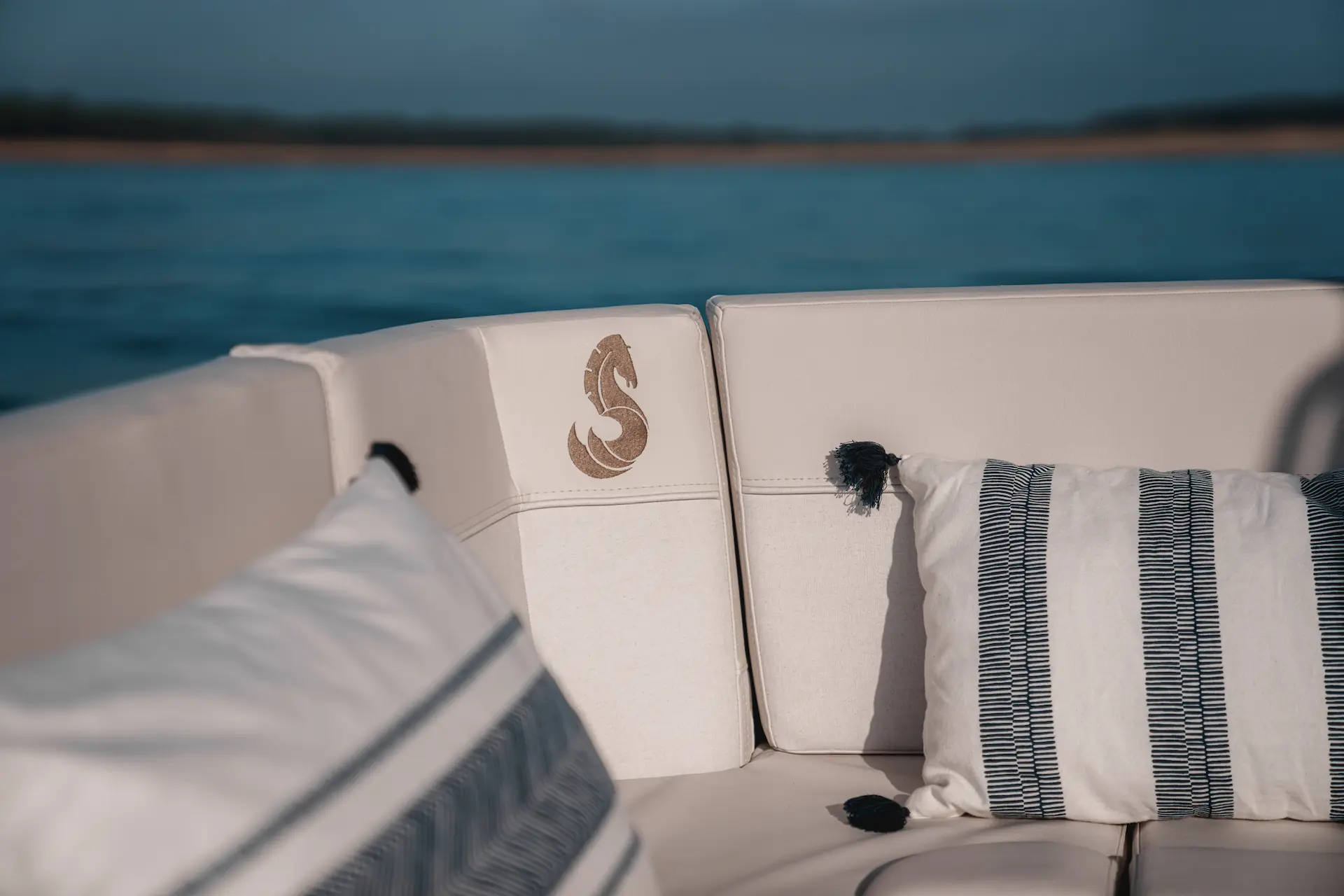 Luxe kussens op een witte zitbank met het zeepaard-logo van Beneteau aan boord van een jacht, met uitzicht op het water.