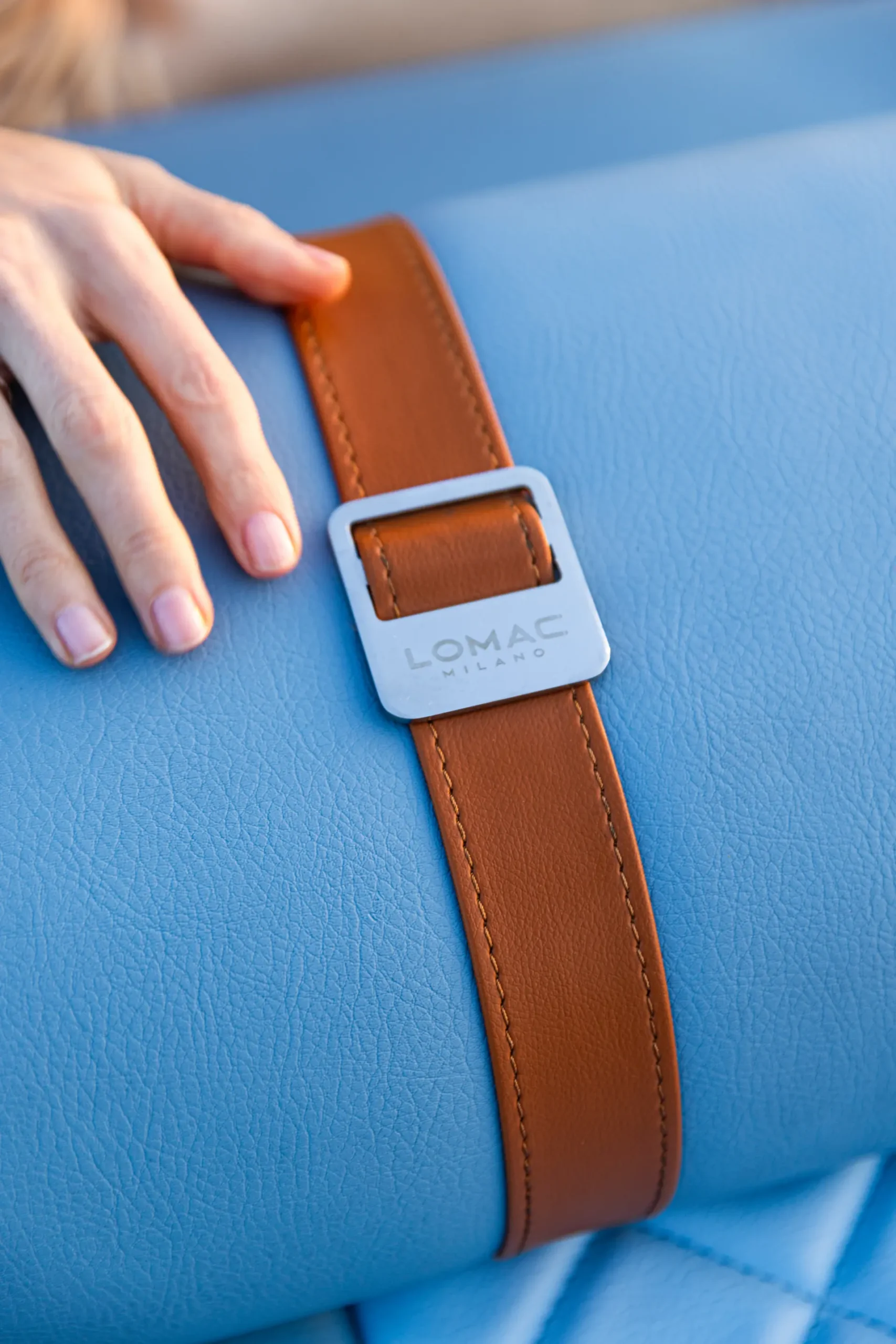 Detail van een cognackleurige leren riem met LOMAC Milano-logo op lichtblauw gestikt leer.